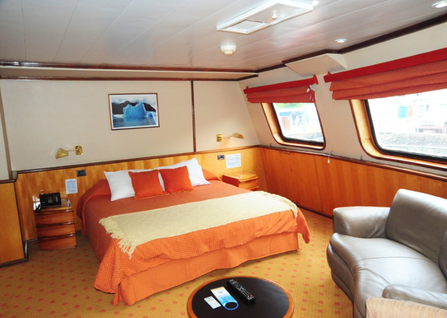 A spacious cruise ship cabin on the Skorpios III Glacier Cruise features a large bed with orange bedding, a gray armchair, and a small round table. Large windows with orange curtains offer an ocean view. The room is decorated with wooden panels and has a picture of an iceberg on the wall.