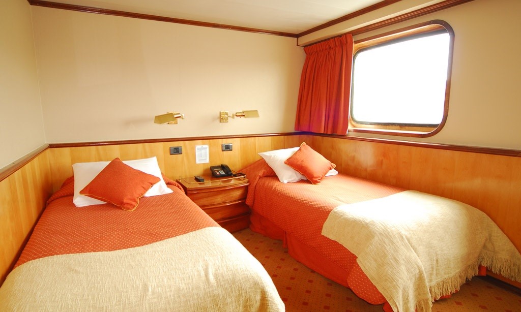 It is a cozy cabin on the Skorpios III Glacier Cruise with two twin beds covered in orange bedding. Each bed has a beige blanket at the foot and an orange throw pillow. A bedside table with a phone and a wall lamp is placed between the beds. A window with orange curtains provides natural light, perfect for viewing Chile's landscapes.