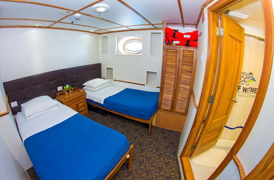 A Tip Top Galapagos cruise cabin with two single beds, each topped with a blue blanket and white pillows. The room features a small wooden nightstand, a porthole window, a life jacket on top of a wooden wardrobe, and a bathroom with a shower enclosed by a curtain.