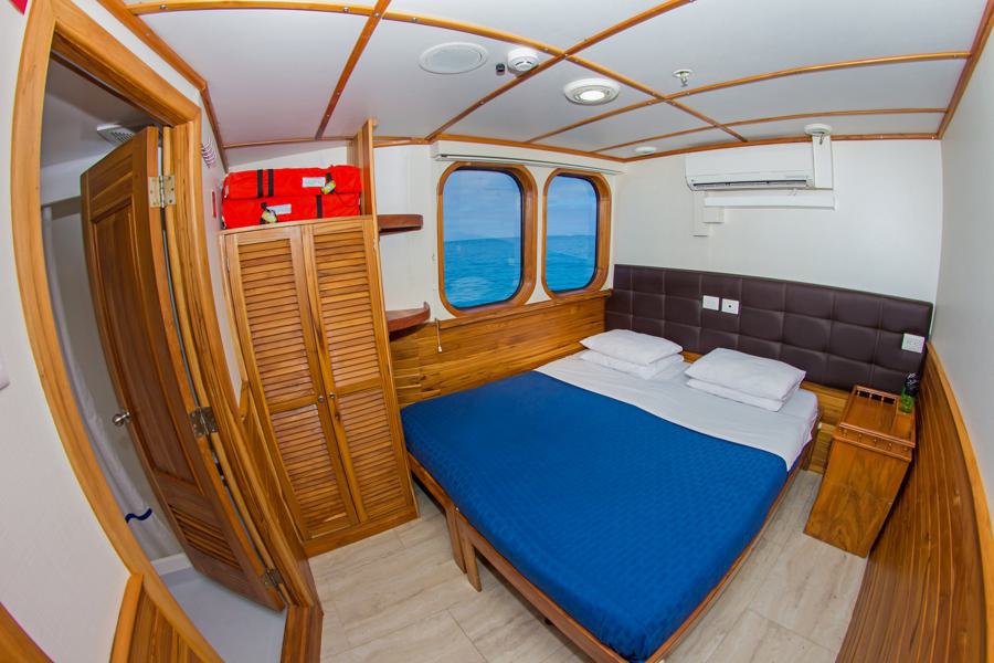 A cozy cabin with a wooden interior features a double bed with a blue blanket, side tables, and large windows offering an ocean view. A wardrobe with louvered doors and life vests on top is next to the bed. The room is well-lit with an overhead light and includes an air conditioner, perfect for your Tip Top Galapagos cruise.