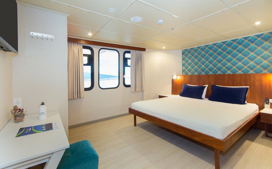 A modern, cozy cabin room with a large bed featuring blue pillows and white linens. The room has two windows with curtains, a small desk with a brochure about the Tip Top Galapagos cruise and a water bottle, a teal chair, and a bedside table. The wall behind the bed has an eye-catching blue geometric pattern.