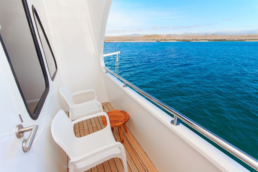 The image shows a boat deck with two white chairs and a small wooden table, overlooking a calm, blue ocean. The deck has a sleek white railing and the horizon in the distance reveals a landmass under a clear blue sky. This serene scene could be from your Tip Top Galapagos cruise adventure.