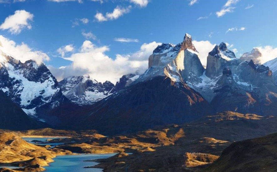 A picturesque mountain landscape featuring snow-capped peaks, rolling valleys, and shimmering blue lakes under a partly cloudy sky in Chile's Torres del Paine. The rugged terrain is highlighted by varying shades of green, brown, and white, creating a dramatic contrast.