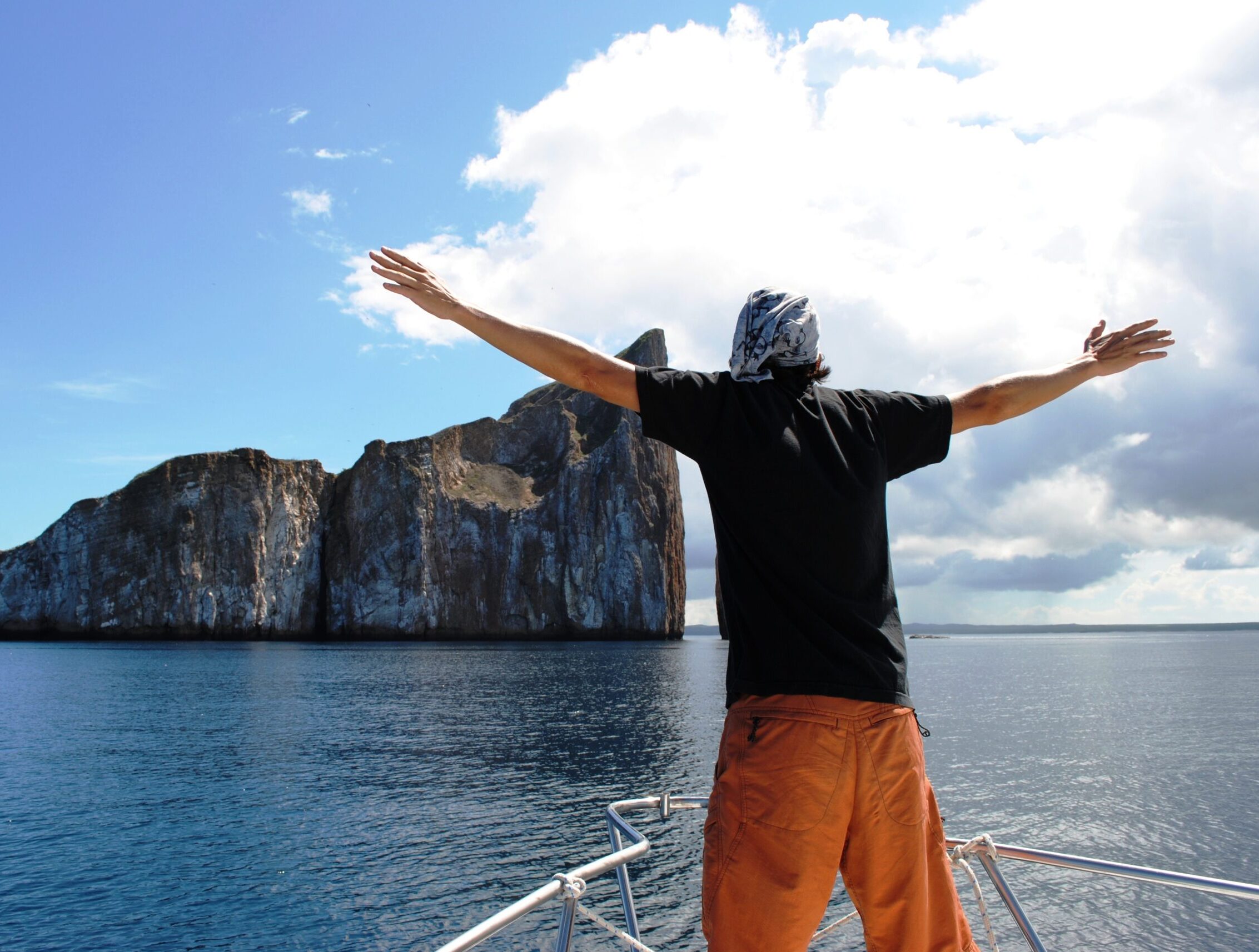 A person stands at the bow of a boat with arms outstretched, facing the ocean. They wear an orange pair of shorts, a black t-shirt, and a headscarf. In the background, a large, rocky island rises from the water under a partly cloudy sky.