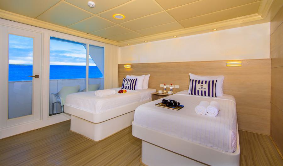 A well-lit cabin on the Treasure of the Galapagos cruise features two single beds adorned with striped pillows and folded towels. A tray with coffee cups sits on one bed. A glass door opens to a balcony with chairs, offering a scenic ocean view under a clear blue sky.