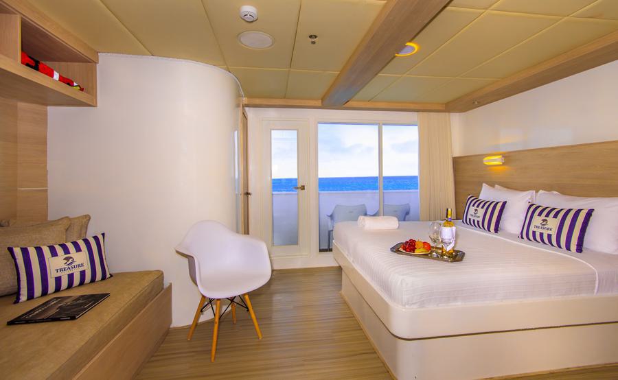 A cozy cruise ship cabin on the Treasure of the Galapagos cruise, featuring a queen-sized bed, white and purple pillows, a sitting area with a beige couch, a white chair, and a table with a tray of fruit and drinks. The room has sliding glass doors opening to an ocean view balcony.