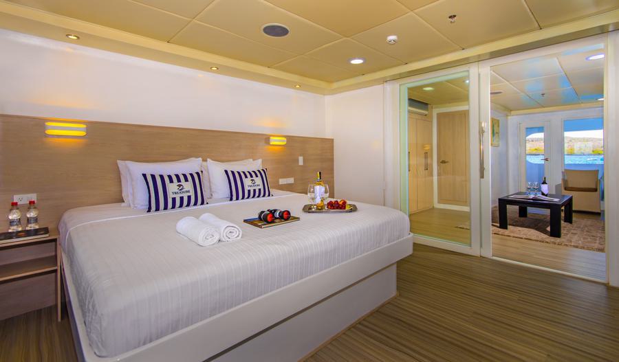 A well-lit, modern hotel room featuring a large bed with white linens and striped pillows. The room includes a nightstand with water bottles, and a small table with fruit and wine. A glass door leads to a cozy sitting area with a view of the outdoors, perfect for those embarking on the Treasure of the Galapagos cruise.