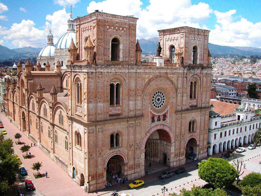 A large, historic cathedral made of reddish-brown stone dominates the image. It features ornate architectural details including multiple arches, towers, and a large rose window. The building is surrounded by a vibrant cityscape with parked cars and people walking nearby.