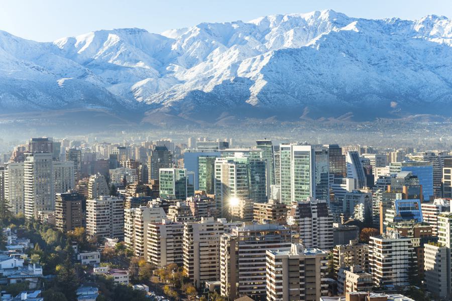 A cityscape of Santiago, Chile, featuring modern high-rise buildings in the foreground and the snow-capped Andes Mountains in the background under a clear blue sky.