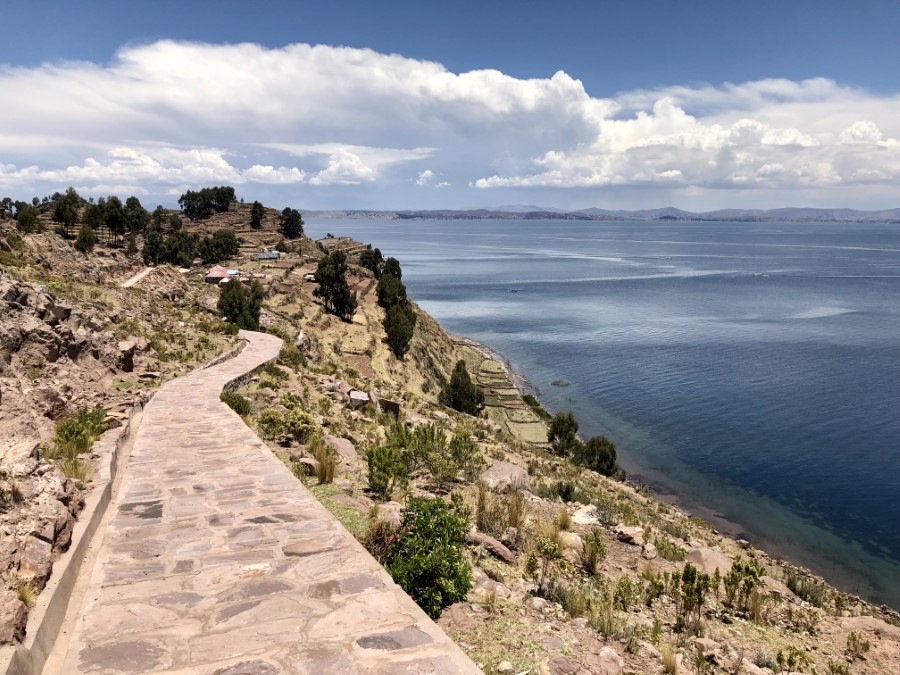 A stone pathway winds along the edge of a hill, overlooking a large blue lake. Terraced fields and trees are visible to the left of the pathway, and distant mountains can be seen across the water under a partly cloudy sky.