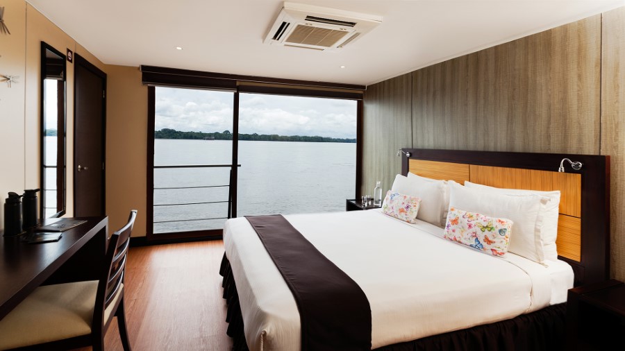 A well-lit hotel room with a large bed adorned with white linens and colorful pillows. The room features a desk and chair, a large window showcasing a scenic lake view, and modern decor with wooden accents. The atmosphere is serene and inviting, much like cabins on the Anakonda Amazon River Cruise ship.