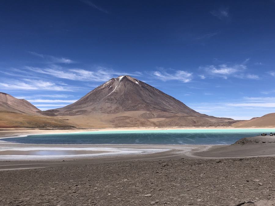 A tranquil scene of a volcano with a snow-capped peak under a clear blue sky. In the foreground, there's a shimmering turquoise lake surrounded by barren, rocky terrain in Chile's Atacama Desert. Sparse vegetation can be seen on the lower slopes of the volcano.