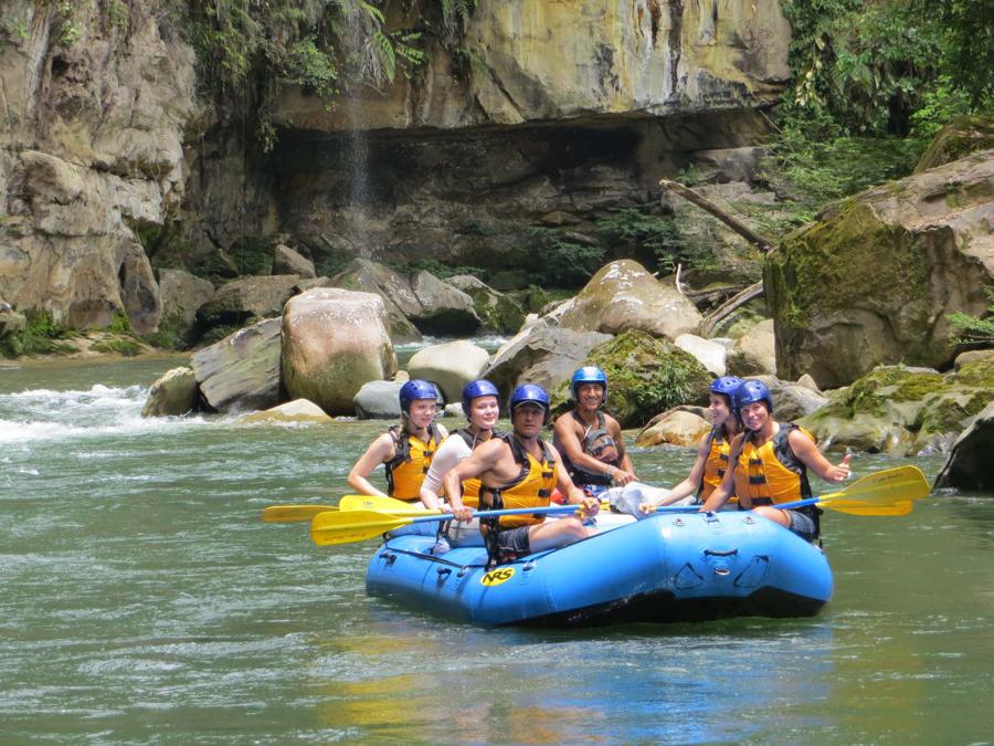 Six people wearing helmets and life vests are seated in a blue inflatable raft with paddles. They are navigating a calm section of a river surrounded by large rocks and lush greenery. A rock formation is visible in the background.