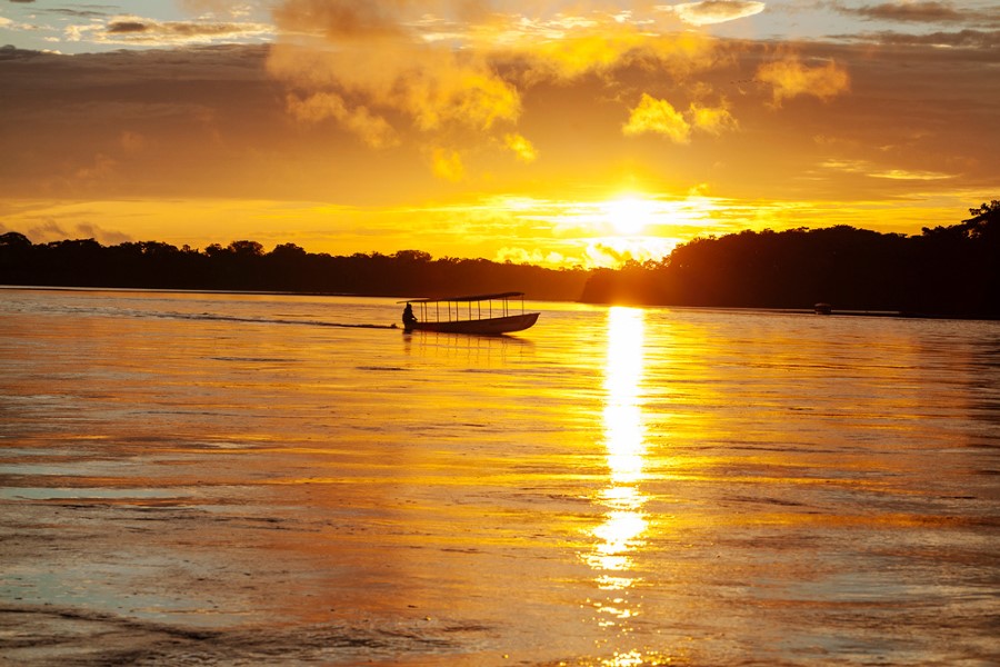 A small boat with a canopy sails on a calm river at sunset. The sky is filled with vibrant orange and yellow hues, reflecting off the water's surface. Silhouetted on the horizon, a tree line creates a serene and picturesque scene.