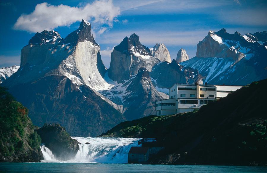 Explora Lodge is a large white building situated on a small peninsula at the base of a mountain range. Snow-capped peaks rise sharply behind it, and a waterfall flows next to the building, emptying into a turquoise lake. The clear sky has some scattered clouds.