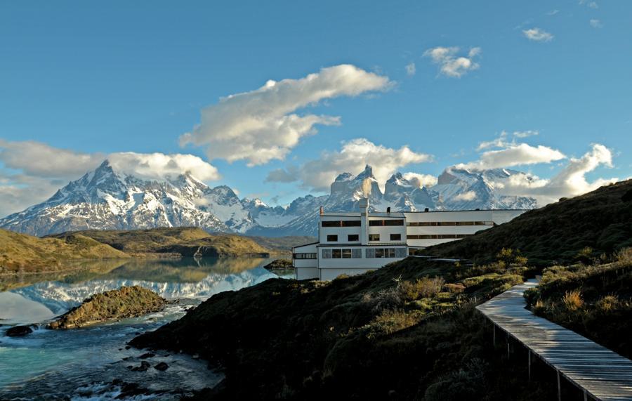 Explora Lodge, a modern white building, stands on the edge of a pristine lake surrounded by lush greenery. Snow-capped mountains and dramatic clouds fill the background. A wooden walkway curves from the foreground to the building, blending natural beauty and architecture.