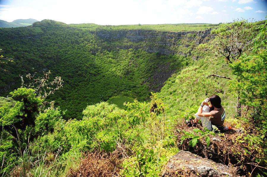 A person sits on the edge of a lush green crater overlooking dense forest vegetation inside the crater. They appear to be holding a child. The sky above is clear and sunny, highlighting the vibrant greenery of the landscape.