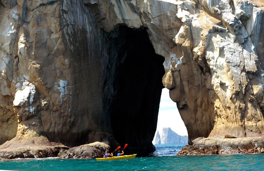 A person in a yellow kayak paddles near a large rocky archway over a body of water. The archway forms a natural tunnel through the rock, with clear blue sky and distant rock formations visible through the opening. The water around the kayak is calm and turquoise.