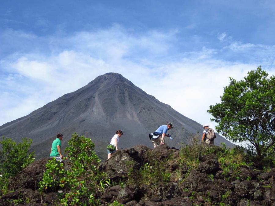 Four people hike up a rocky path with lush green vegetation, with a large, cone-shaped volcano towering in the background under a partly cloudy blue sky.