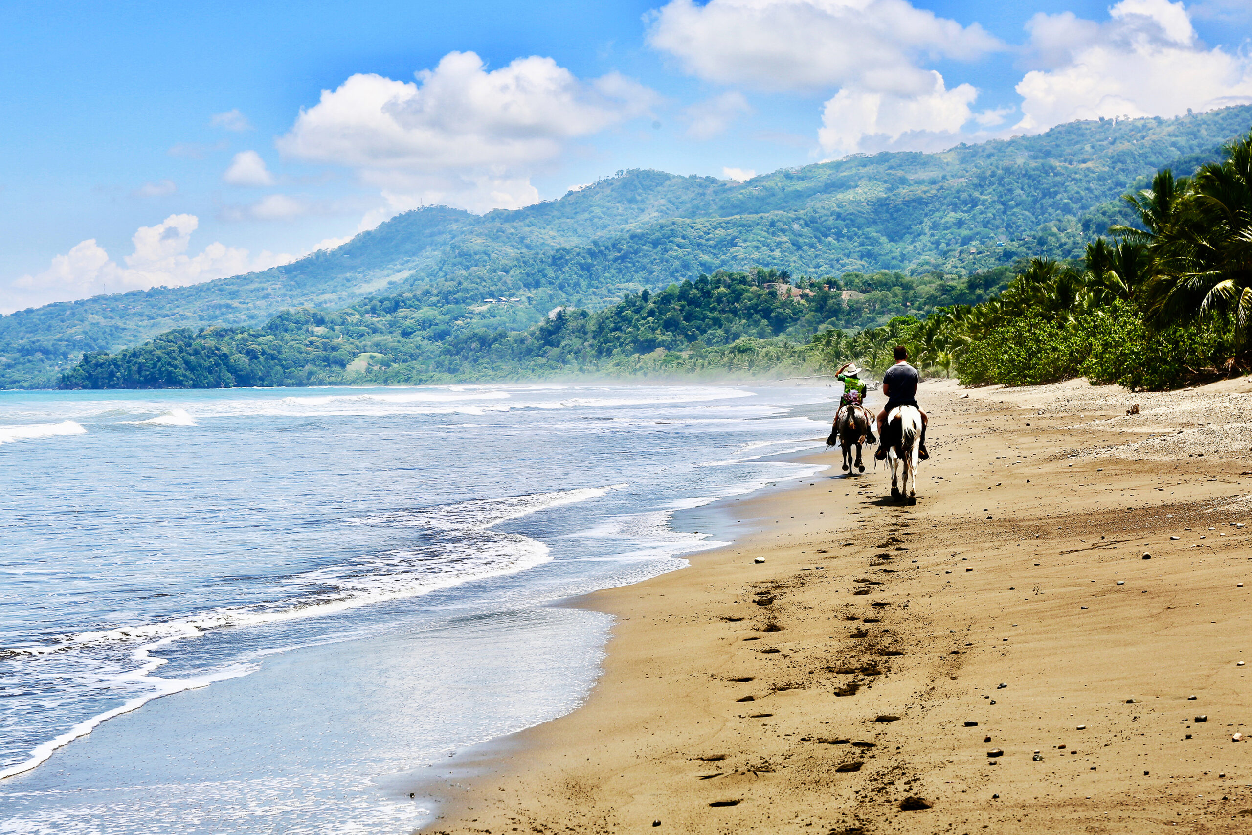 Two people ride horses along a sandy beach with footprints trailing behind them. The serene beach is bordered by lush green hills and trees, under a bright blue sky with scattered white clouds. The calm ocean waves gently lap the shore.