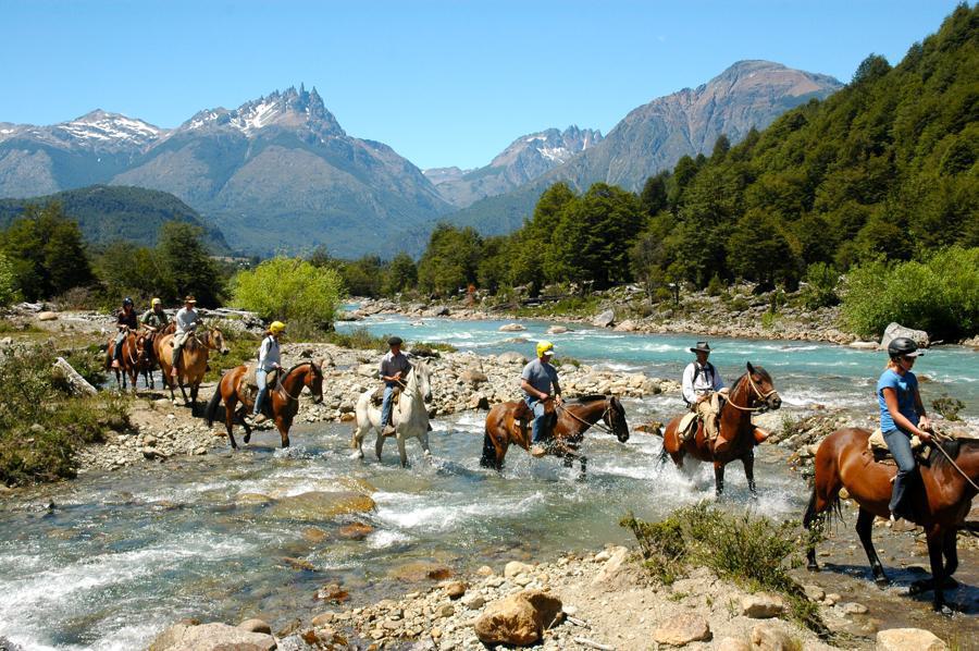 A group of people rides horses across a shallow river in a scenic mountain landscape. Snow-capped peaks and lush green trees fill the background. The weather is clear and sunny, suggesting a pleasant day for horseback riding amidst nature.