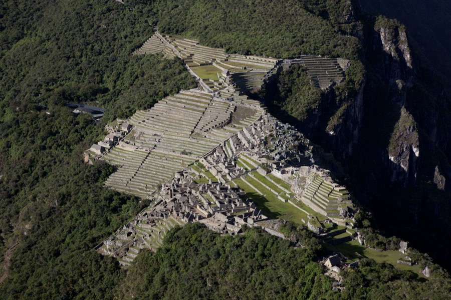 Aerial view of the ancient Incan city of Machu Picchu. Stone terraces cascade down the mountain, with lush green vegetation surrounding the historic stone structures. The mountains and dense forest encase the ruins, highlighting the remote location.