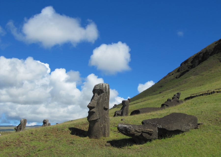 Several moai statues of varying sizes are partially buried in the grassy hillside of Rano Raraku on Easter Island, with a clear blue sky and scattered white clouds in the background. The largest statue in the foreground features a prominent, distinct facial profile.