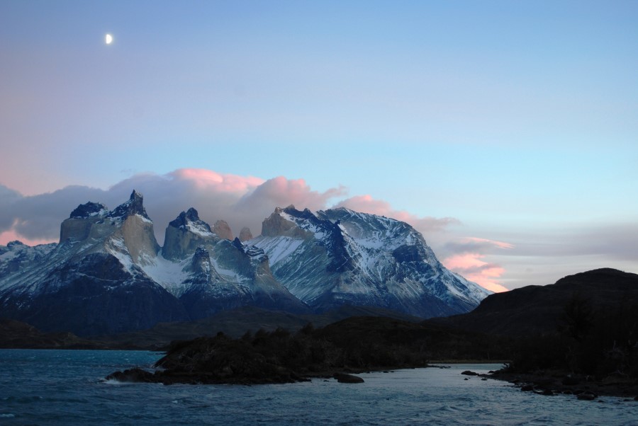 A stunning landscape of the Torres del Paine mountain range in Patagonia, Chile, captured during sunset with a crescent moon in the sky. Snow-capped peaks are highlighted by soft, pink-tinged clouds. A serene lake and rugged foreground add depth to the scene.