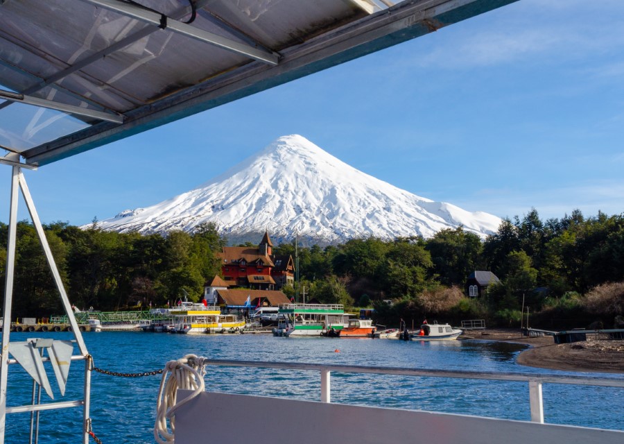 A scenic view of a snow-capped volcano with a tranquil lake in the foreground. Boats are docked at the shore, with a charming house and lush green trees surrounding the area, all captured from the perspective of a boat on the lake.