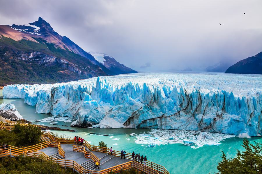 A large glacier meets bright turquoise water surrounded by lush green hills and high mountains under a cloudy sky. A wooden viewing platform with railings and pathways extends into the foreground, where a group of people are looking at the Perito Moreno Glacier in Argentina.