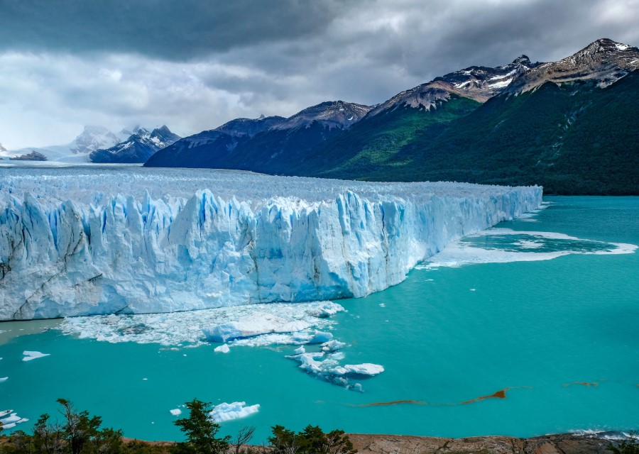         Image of the Perito Moreno Glacier extending into a turquoise lake, surrounded by rugged mountains with patches of snow and verdant forest. The sky is partly cloudy with a mix of dark stormy and clear areas. A few icebergs are floating near the glacier's edge in Argentina.