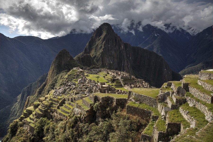 A view of Machu Picchu, the ancient Incan city nestled in the Andes Mountains, under a cloudy sky. The stone structures and terraces of the ruins are prominently visible, with lush green mountains surrounding the site.