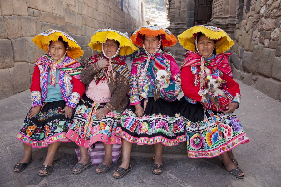 Four women sit on a stone bench, dressed in traditional, colorful Andean clothing. They wear intricately embroidered skirts, shawls, and yellow hats, each holding small, white furry animals. The backdrop features a cobblestone street with stone walls.