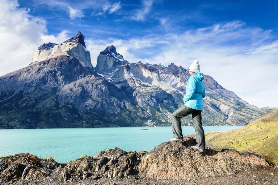 A person wearing a blue jacket, gray pants, and a white beanie stands on a rocky outcrop, gazing at a turquoise lake and the towering, jagged mountains under a partly cloudy sky. The scene is set in the rugged mountainous landscape of Torres del Paine, Chile.