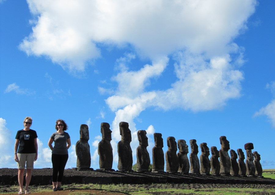 Two people stand on a grassy area next to a row of large, ancient stone statues known as moai, under a bright blue sky with scattered clouds on Easter Island. The statues are lined up in a row facing inward, and the people appear to be tourists.