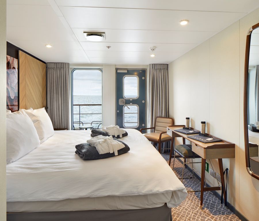 A neatly made bed with white linens and a bathrobe is in the center of a modern cruise ship cabin. The room features a large window with an ocean view, a small desk and chair, a wall mirror, and neutral-colored decor. Ideal for relaxing after a day on the Magellan Explorer during your Antarctica expedition.