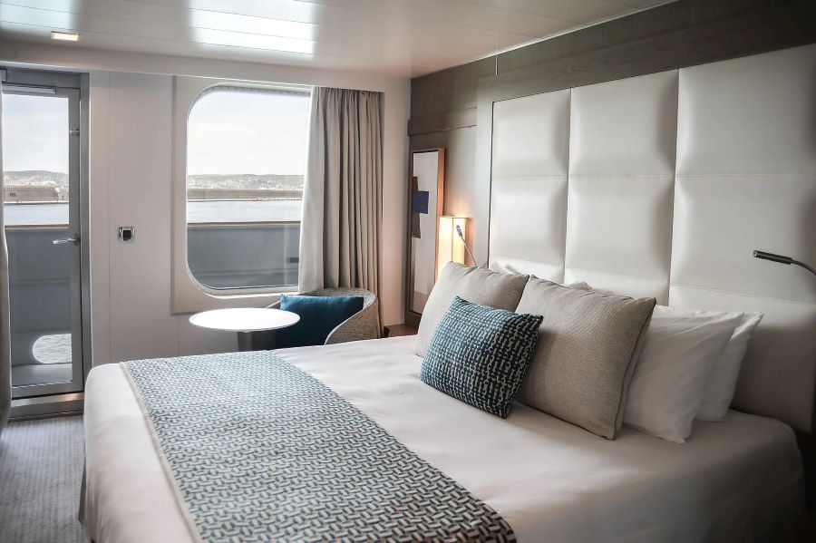 A modern cruise ship cabin with a large bed adorned with white and patterned blue linens. The room features a window, a small table with a chair next to it, and curtains drawn to one side. The sleek décor in neutral tones complements the view of your Ponant Antarctica expedition from the balcony door.