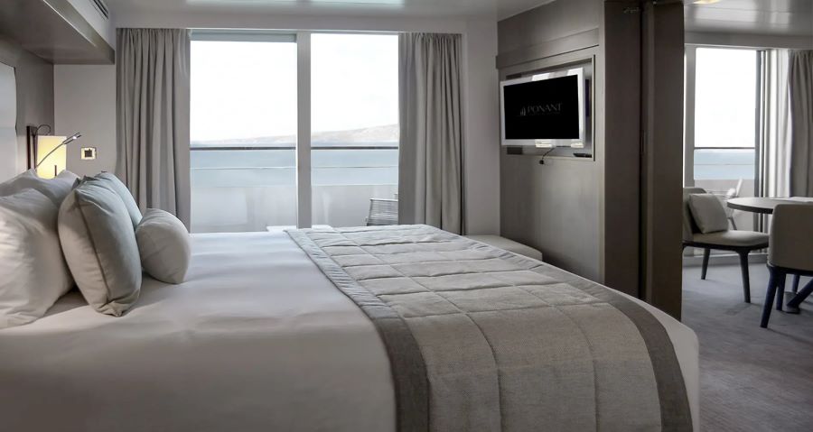 A modern hotel room with a king-sized bed, white and grey bedding, and floor-to-ceiling windows showing an outdoor balcony and scenic view. A television is mounted on the wall, and a sitting area with a table and chairs is visible in the background, perfect for planning your Ponant Antarctica expedition.