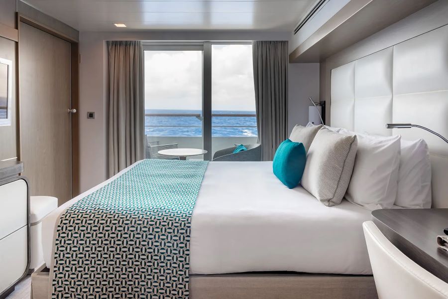 A modern hotel room with a neatly made bed featuring a geometric patterned blanket and blue accent pillows. The room boasts a large window or sliding door with curtains, leading to a balcony with an ocean view reminiscent of a Ponant Antarctica expedition. Minimalistic furniture and neutral colors create a clean look.