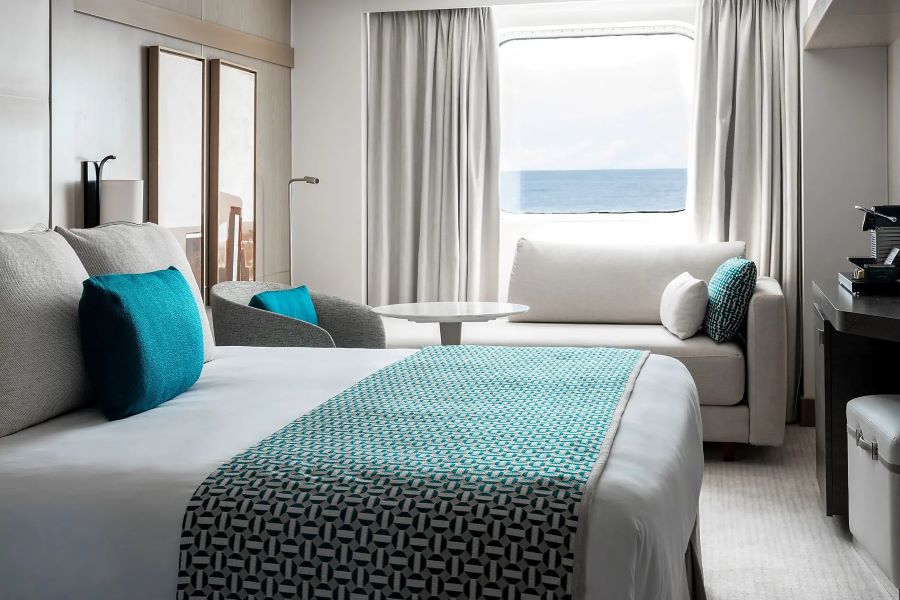 A modern, well-lit hotel room with a large bed adorned with blue and white geometric patterned bedding and blue cushions. The room features a small lounge area with a sofa, armchair, coffee table, and a large window offering an ocean view similar to that of a Ponant Antarctica expedition.