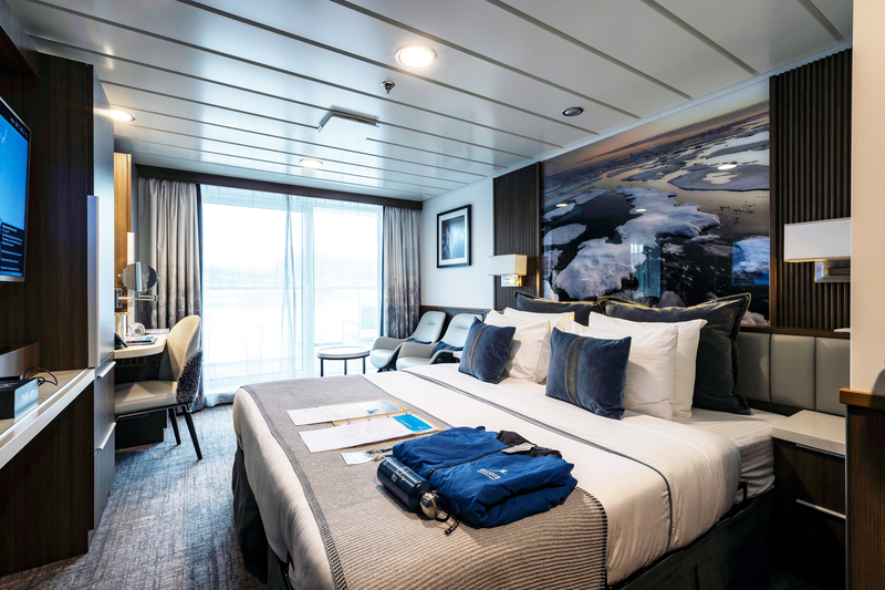 A modern hotel room with a large bed adorned with white and dark blue bedding. The room features a flat-screen TV, desk, two chairs, and a large window with sheer curtains. Folded blue clothes, headphones, and papers from the Sylvia Earle Antarctica expedition are neatly placed on the bed.