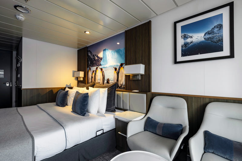 A cozy, modern bedroom featuring a comfortable double bed with multiple pillows, a large wall art piece of penguins by an iceberg from the Sylvia Earle Antarctica expedition, two white and gray chairs, and framed photography of icy landscapes. Warm lighting enhances the soothing, minimalist decor.