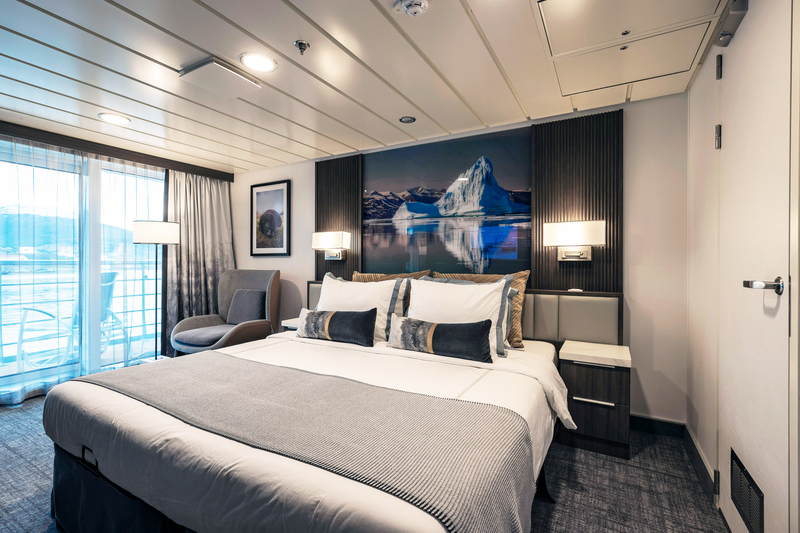 A modern hotel room with a large bed adorned with white sheets and grey pillows. The room features a photo from the Sylvia Earle Antarctica expedition on the wall, two bedside tables with lamps, an armchair, and a balcony with sheer curtains that lets in natural light.