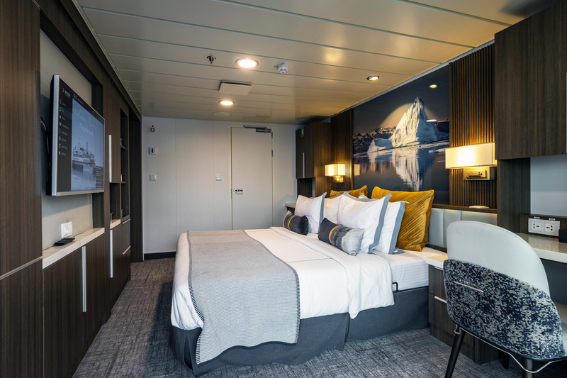 A modern hotel room with a queen-sized bed, decorated with white and gray bedding and gold accent pillows. A large scenic art piece of a mountain from Sylvia Earle's Antarctica expedition hangs above the bed. The room also features a wall-mounted TV, work desk, built-in storage, soft lighting, and carpeted floors.