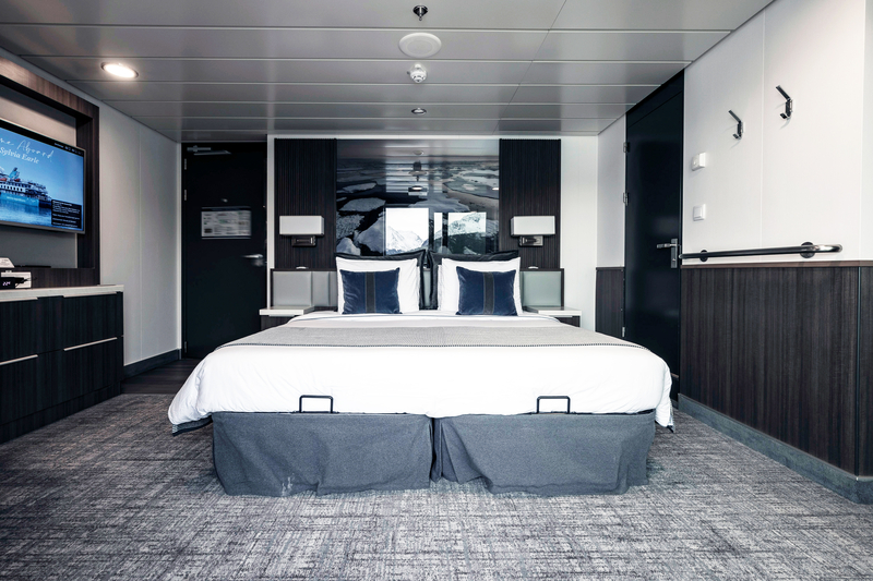 A modern hotel room with a large bed centered against a wall, featuring a dark headboard and white bedding with dark accent pillows. Wall-mounted TV on the left and a dark door to the left side of the bed. The decor draws inspiration from Sylvia Earle's Antarctica expedition, with contemporary dark wood tones and gray carpet.