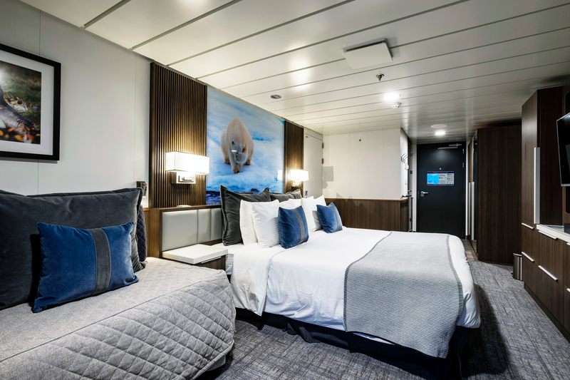 A modern, well-lit hotel room features a large bed with white linens and blue accent pillows. To one side, there is a cozy daybed with gray bedding. The walls are adorned with contemporary artwork inspired by Sylvia Earle's Antarctica expedition, and there is a dark-toned wardrobe opposite the bed.