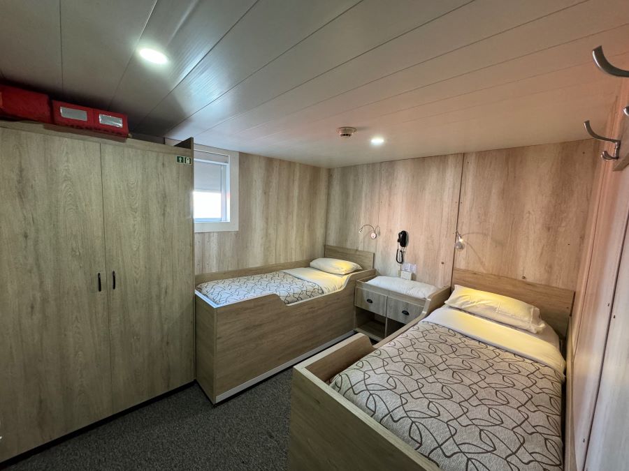 A cozy room featuring twin beds with wooden frames and patterned bedspreads. The room has wood-paneled walls, a small window, wall-mounted reading lights, and a bedside table. On the left is a large wooden wardrobe, evoking the warmth needed after an Ushuaia Antarctica expedition.