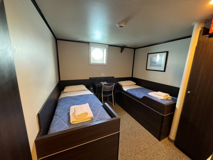 A small, well-lit cabin with two single beds and a single porthole. Each bed has blue bedding and a neatly folded towel. There’s a small desk with a chair beneath the porthole, perfect for planning your Ushuaia Antarctica expedition. A framed picture adorns the wall, complementing the room's minimalistic decor.