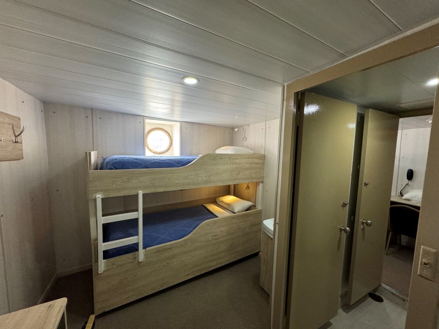 A compact cabin featuring wooden bunk beds with blue bedding and white pillows is perfect for an Ushuaia Antarctica expedition. Nearby, there's an open door leading to a small, mirror-lined bathroom. A small, round porthole window above the top bunk allows light into the room. Walls are paneled in light wood.