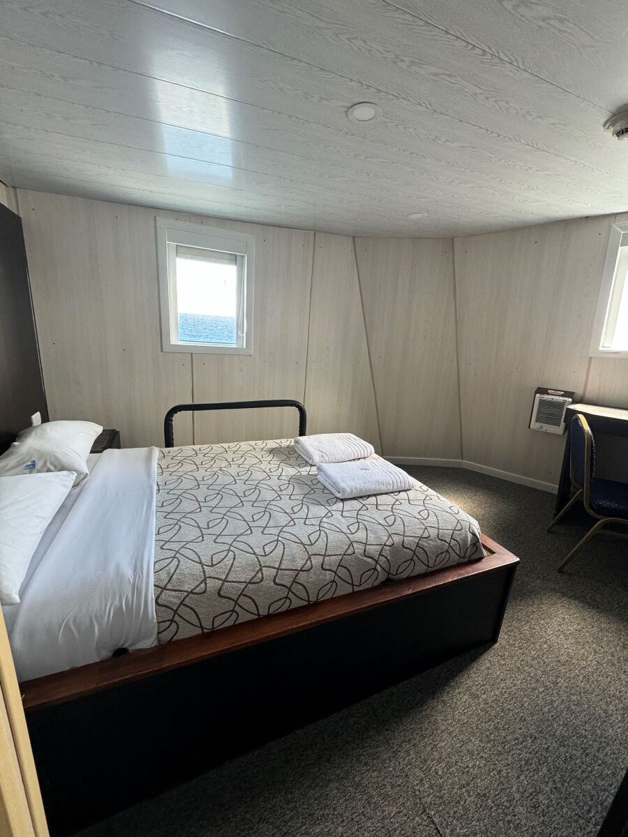 A small room with wood-paneled walls features a double bed with a white duvet and patterned blanket. Natural light enters through two small windows. The room also contains a chair and desk in the corner, perfect for planning your next Ushuaia Antarctica expedition. Towels are neatly folded on the bed.